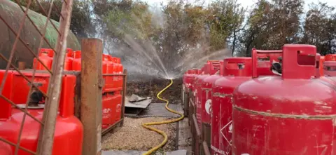 HIWFRS Water spraying out from a fire crew's hose in front of gas cylinders seen lined up in rows.