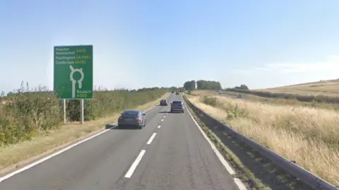 Cars travel along a dual carriageway with long grass and fields either side of the road, and a sign indicating an upcoming roundabout.