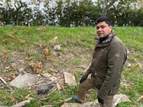 Dr Muhammad Ali is standing in front of a grassy embankment with some exposed earth. There are chunks of rubble visible on the side of the bank, including what appears to be concrete and asphalt.