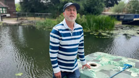 Man with grey beard in navy cap wearing white and blue-striped rugby style shirt stood on the edge of an abandoned boat on a canal