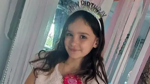 Family Aria smiles at the camera in front of a doorway filled with pink streamers. She has long brown wavy hair and is wearing a pink dress and shrug. She has a bright pink birthday badge on, and a sparkly headband which reads "HAPPY BIRTHDAY".