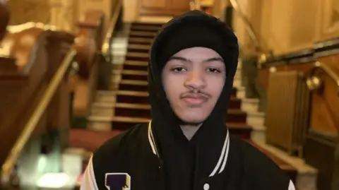 Ayshea Buksh/BBC Tryelle Johnson, wearing a black jacket with white lines on the collar and a black hat, is looking straight at the camera. Stairs from the theatre can be seen in the background