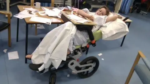 PA Images Jamilla Blake as a child in a hospital ward in her reclining wheelchair. She is wearing a hospital gown and smiling at the camera.
