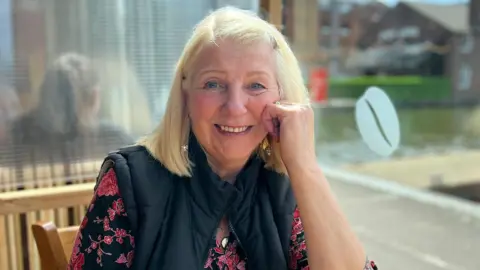 Pamela Dumbleton A woman sits at a cafe table with sunlight streaming through the window beside her. She is wearing a patterned red and black top with a dark padded gilet. Outside the window, a canal and buildings are visible under a bright sky.
