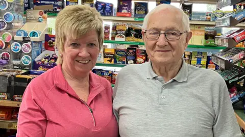 BBC/Dan Sambell Teresa is wearing a pink top and had short blonde hair. She is standing next to Patrick who is wearing a grey polo shirt and has grey hair. They are standing in front of the counter of the shop.