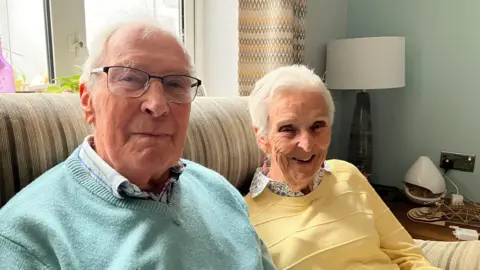 Amy Holmes/BBC Kenneth and Elvina Calder smile at the camera as they sit on a sofa that is positioned in front of a window. Kenneth is wearing a plaid shirt, turquoise jumper and glasses. Elvina is wearing a yellow jumper, shirt and pink earrings. Behind Elvina is a lamp and curtains. 