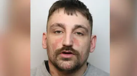 Huckvale stares at the camera with a blank look on his face. He has brown hair, stubble, and a thick moustache and eyebrows. 