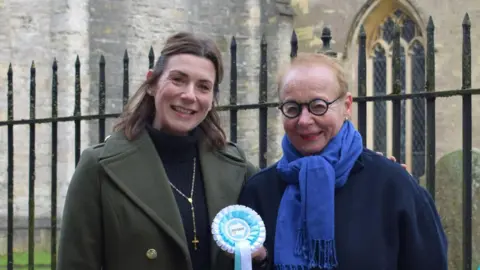 two women standing outside a church railing with a light blue rosette