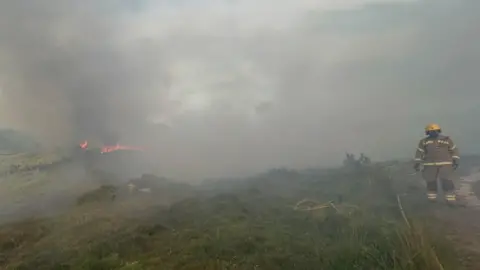 A firefighter stands on a track in a smoke-filled landscape with a fire burning in the distance. 