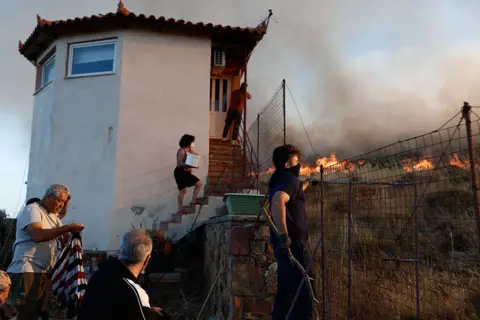 Getty Images Residents try to contain a wildfire near a small building outside the town of Chios island in Greece, June 22, 2025. One lady is holding a bucket of water.