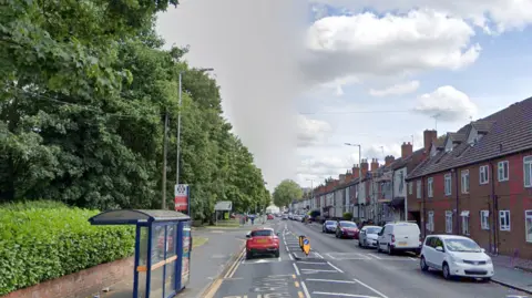 Google A bus stop is on the left of the road in front of a row of trees with houses on the other side and cars parked outside.