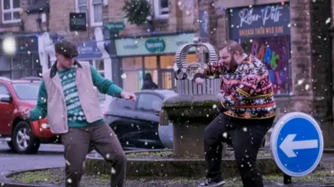 Reel Productions Two men in Christmas jumpers are dancing on a roundabout. The man on the right has short dark hair, a beard, black trousers, trainers and a colourful jumper which includes snowmen, candy canes and reindeer. The man on the left is wearing a green Christmas jumper covered partially by a cream bodywarmer, brown trousers and a brown flat cap. There is a blue sign with a white arrow on the roundabout and a sculpture of a crown. A black car and a red car are driving around the roundabout with shops in the background. The image has been edited so that it looks like it is snowing.