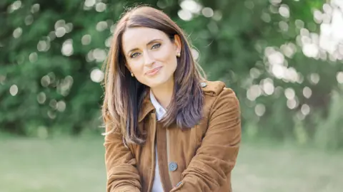 A glamorous image of Anne taken at the Royal Highland Show - she wears a tan suede jacket and her hair is long. She is smiling and there are trees and grass behind her.