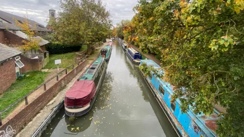 Chris Andrews Publications A view of the Mount Place bridge of the Oxford Canal during the Jericho Oktoberfest on 17 October. Several boats are lined up along the bank and trees. It is cloudy.