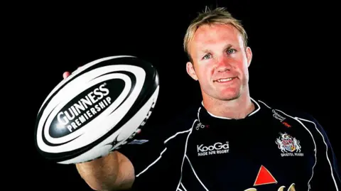 Matt Salter holding the black and white Guiness Premiership rugby ball t the camera. He is wearing a dark kit with red white and gold accents.
He has blond hair and blue eyes and is smiling at the camera.