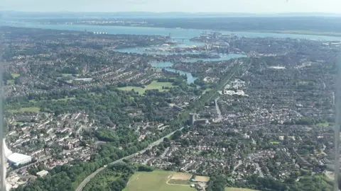 An aerial view of a residential area, with streets of houses spread out from parkland, leading to a river which flows in to the sea