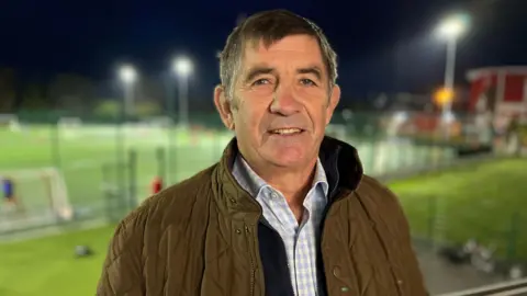Philip Wilkinson stands above a sports pitch, which is lit by floodlights on a dark night, with his back to it. He is wearing a brown quilted coat, a dark fleece, and a light blue and white checked shirt. He has dark grey hair and is smiling.