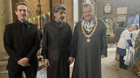 Three men stand in a line smiling at the camera. They are standing in the cathedral, with the cathedral walls seen in the background. The men are wearing different dress - the man on the far left wears a black suit and tie. The man in the middle is wearing a black suit with a collarless waistcoat, and the man on the far left wears a long, grey robe with a golden mayor-like livery collar around his neck. 