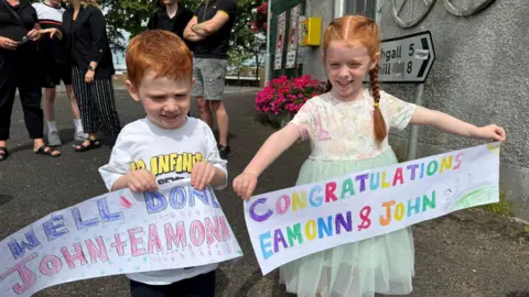 Two red haired chidren, a boy and a girl, hold colourful handmade banners, their greeting, all in uppercase letters, filled in bold colours. Behind them adults stand around talking.