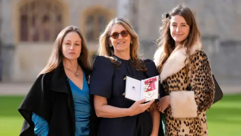 Andrew Matthews/PA Wire A woman wearing a navy dress with balloon sleeves, stands outside whilst holding an award in her hands. A woman wearing a leopard print coat is to her left, whilst a woman wearing a blue silk dress and shawl stands to her right.