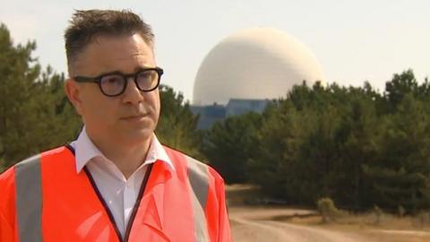 Tom McGarry with short dark hair wearing an orange hi-vis in the shadow of Sizewell nuclear plant