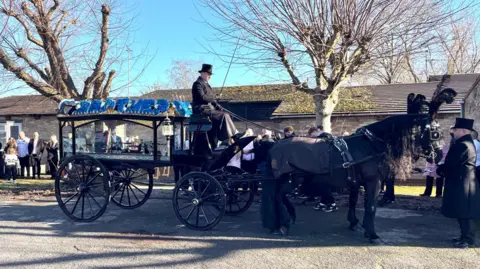 Stephen Huntley/BBC A stationary funeral carriage drawn by two black horses is attended by two men in black top hats, while groups of people can be seen in the background.
