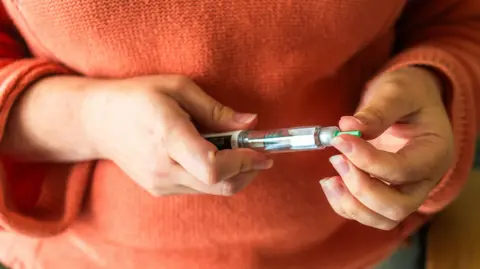 Woman wearing an orange jumper holding a weight loss injection.