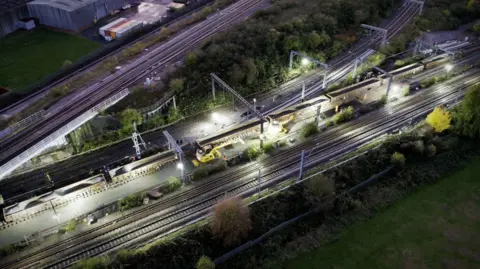A number of train tracks pictured at dusk, with lights and railway workers, wearing orange. A number of buildings can also be seen in the background.