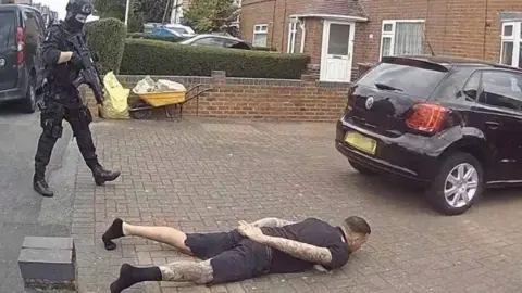 Nottinghamshire Police Man with tattoos and dark clothing lying down on floor as armed police officer stands over him.