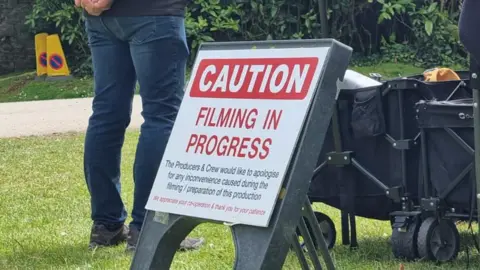 A film crew member wearing blue jeans and black boots stands on a lawn. A sign saying "caution - filming in progress" is on the lawn along with two trolleys filled with equipment.