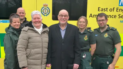 Tony Wright is in the middle and is wearing a black long coat and blue shirt. His wife is stood to the left of him and has short grey hair and is wearing a beige coat and white scarf. Four emergency services staff are stood around them, with three of them wearing paramedic uniform. 