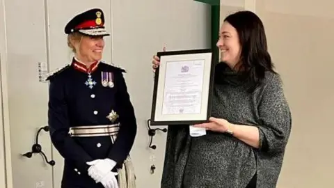 Sunday Centre King's representative Dame Hilary Chapman, the Lord-Lieutenant for South Yorkshire, wearing a navy uniform with medals, military cap and white gloves, presents the framed award certificate to the Sunday Centre's chair Kelly Lingard wearing a grey top