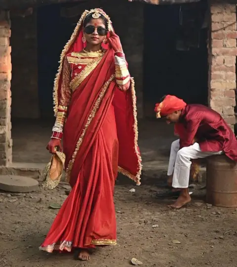 Innovation for Change A young girl modeling in a viral fashion show walks down the street wearing colorful red clothes, jewelry, and sunglasses, while a man sits behind her on the roadside, putting on his shoes. I am.