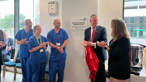 Health staff and MP Luke Pollard stand inside the new centre next to a plaque. There are large windows behind them. The group of people are clapping their hands together and are smiling at one another.