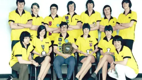 A group of 13 people dressed in yellow t-shirts posing with a trophy in the 1970s. 