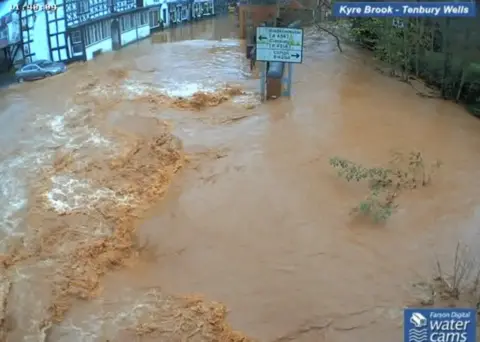 Farson Digital Watercams Brown water rises in a town centre and reaches halfway up a single street sign in the middle of the flow, while water also laps at the foot of buildings in the distance