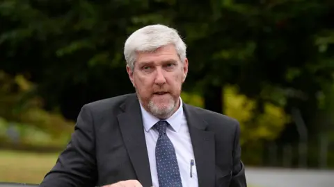 PA Media John O'Dowd dressed in a grey suit with a grey beard and a pen in his chest pocket. He has white hair and a white beard and is stood in front of trees.