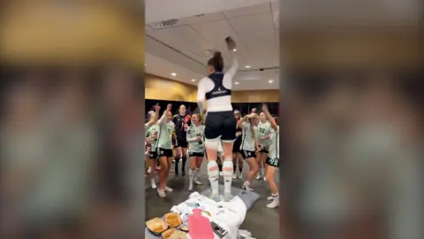 A female football team in a changing room. They are gathered around a footballer who is standing on the bench and cheering her. 