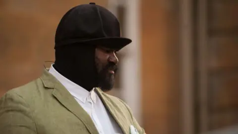 Getty Images Gregory Porter performing at the palace wears a cap and snood like item. He is wearing a green blazer over a white button-up shirt. Out of focus behind him is the palace.