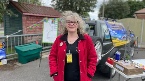 A woman in a red coat is standing in a car park next to a model of a pylon and in front of a car draped in protest banners