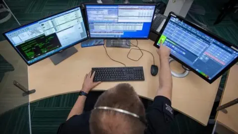 A view from above and behind of a man with brown hair wearing headphones at a desk. He is touching the screen of one of three monitors with his right index fingertip. All three monitors are displaying different files and systems. 