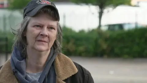 Debbie with shoulder length grey hair wearing a grey baseball cap, grey hoodie and t-shirt and brown faux fur-lined coat stands in a street in Salford. She looks impassive.