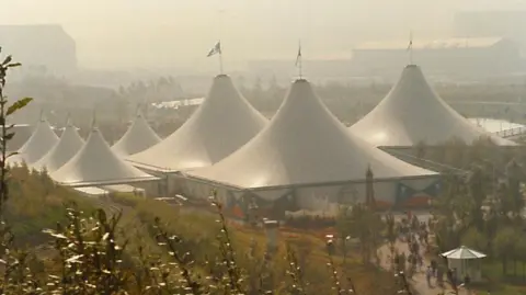 Potteries Museum and Art Gallery A series of large white tents - three very large and four smaller - are positioned among a lawned area which also includes flowerbeds. There is a train track running alongside the left hand side of the image. In the distance are some other buildings which are obscured by the hazy atmosphere.