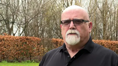 Gary Palmer gives a TV interview in a grassed area in front of a hedge. He is bald on top, with a white goatee beard, and wears an open-necked black shirt and glasses.