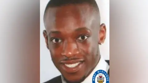 Policía de West Midlands. Un hombre con cabello negro corto, bigote y barba recortados viste una camisa blanca con cuello y una chaqueta negra. Mira a la cámara, frente a un fondo blanco.