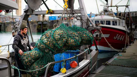 PA Media A fisherman is unloading nets of scallops from his boat onto the quay in St Helier harbour. The green nets are packed with scallops. He is wearing a woolly hat and a hoodie and has black hair and a black beard. 