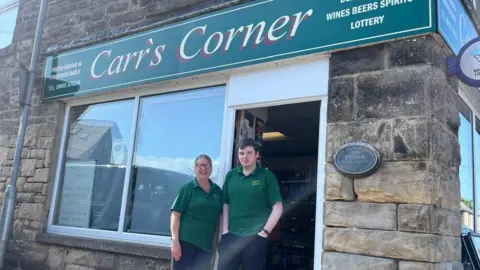 James Robinson/NCJ Media Trudy Long and Thomas Fox are stood in their butchers uniforms outside of Carr's Corner. It is a stone building with a green sign with above the door reading "Fresh bread and produce daily".