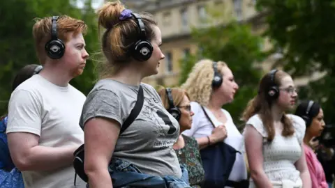 AFP via Getty Images A group of visitors listen on their headsets to a our guide during a visit to discover the sets of the Netflix series "Bridgerton", in Bath. The one man and five women wear headsets and a Regency era grand building sits behind them.