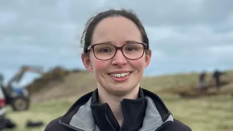 A woman with dark hair tied back and black glasses smiles at the camera. She has a black coat and a black fleece on. Behind her the background is blurred but there is a blue tractor, a black pick-up truck and volunteers working.