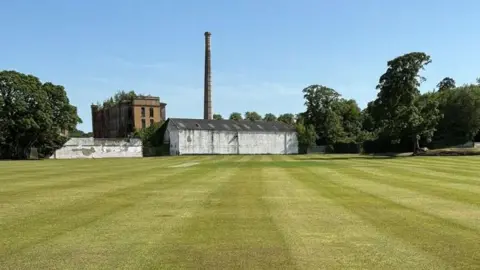 Sion Mills Cricket Club A green grassed grass cricket field stretches towards an old industrial building with a high chimney stack. The sky is blue. A number of trees are on either side of the building.